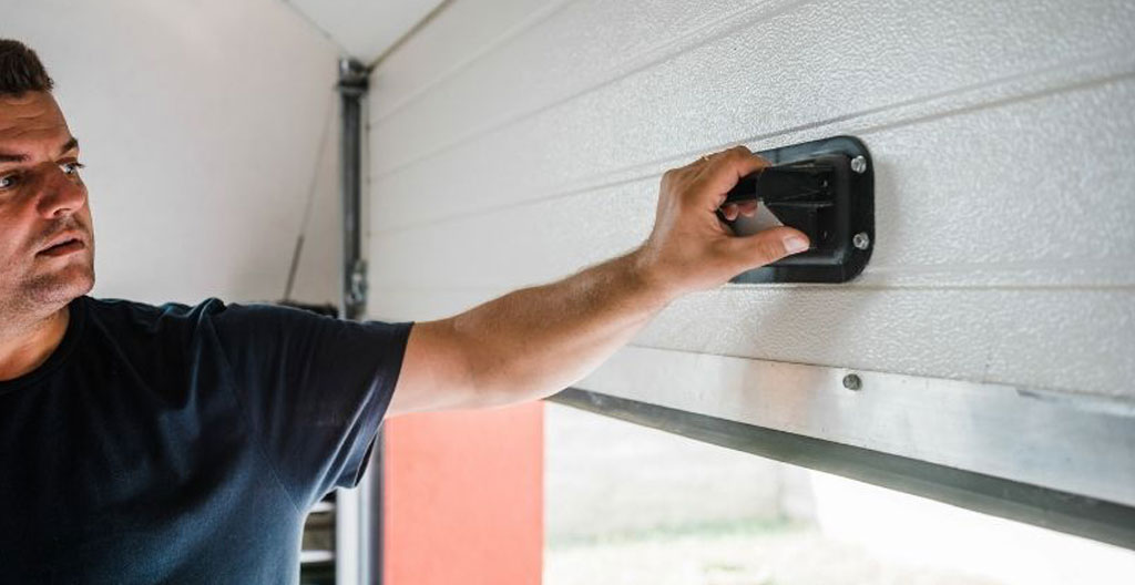 Technicien en portes de garage accroupi inspectant le coupe-froid au bas d'une porte, lampe de poche dirigée vers le joint